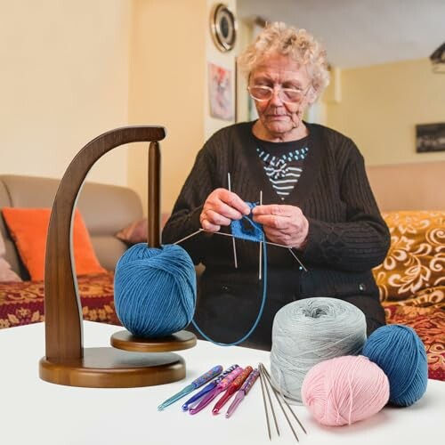 Elderly woman knitting with blue yarn on a table with multiple yarn balls and crochet hooks.